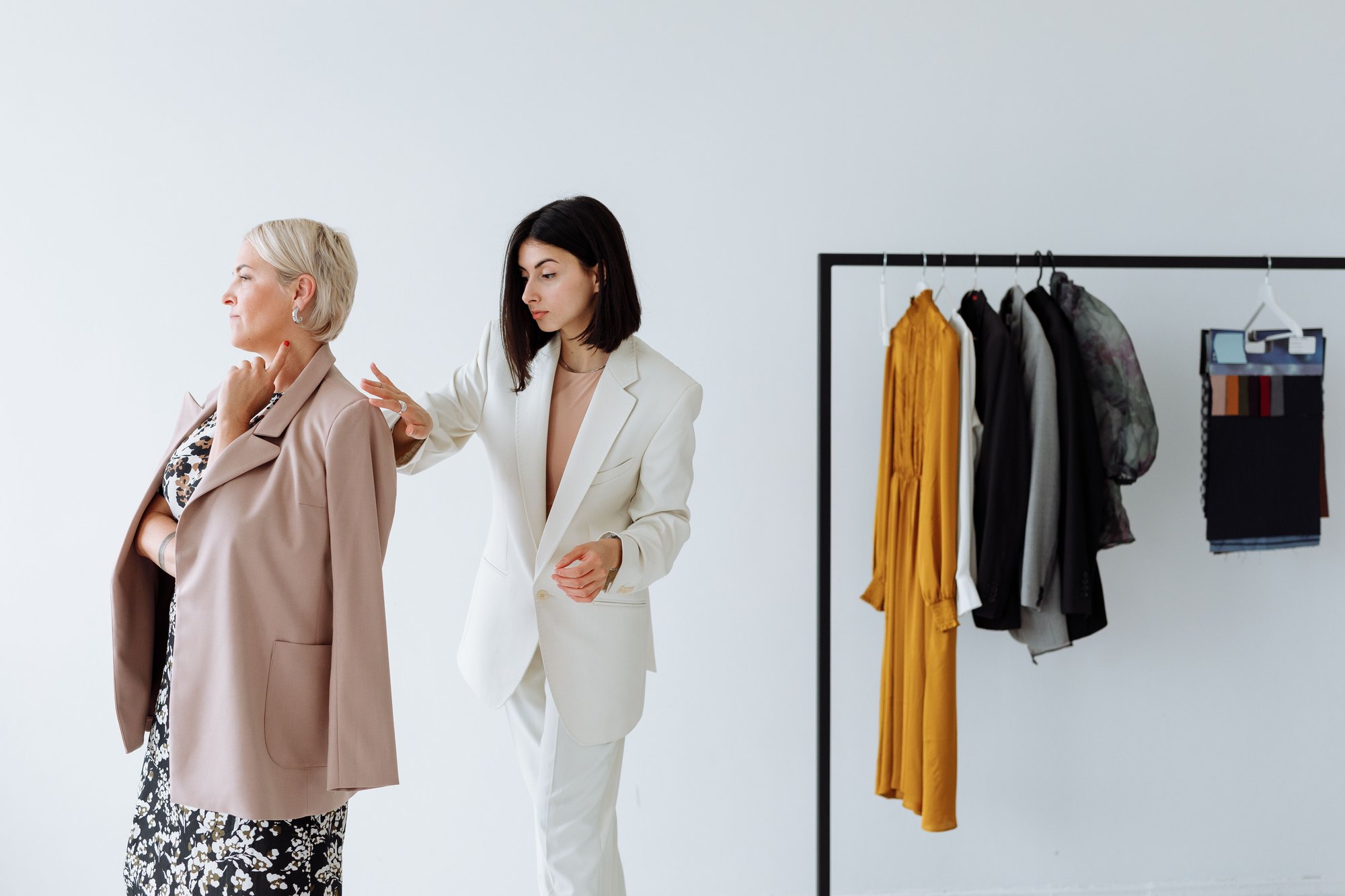 A stylist fitting a jacket for a client beside a rail of garments